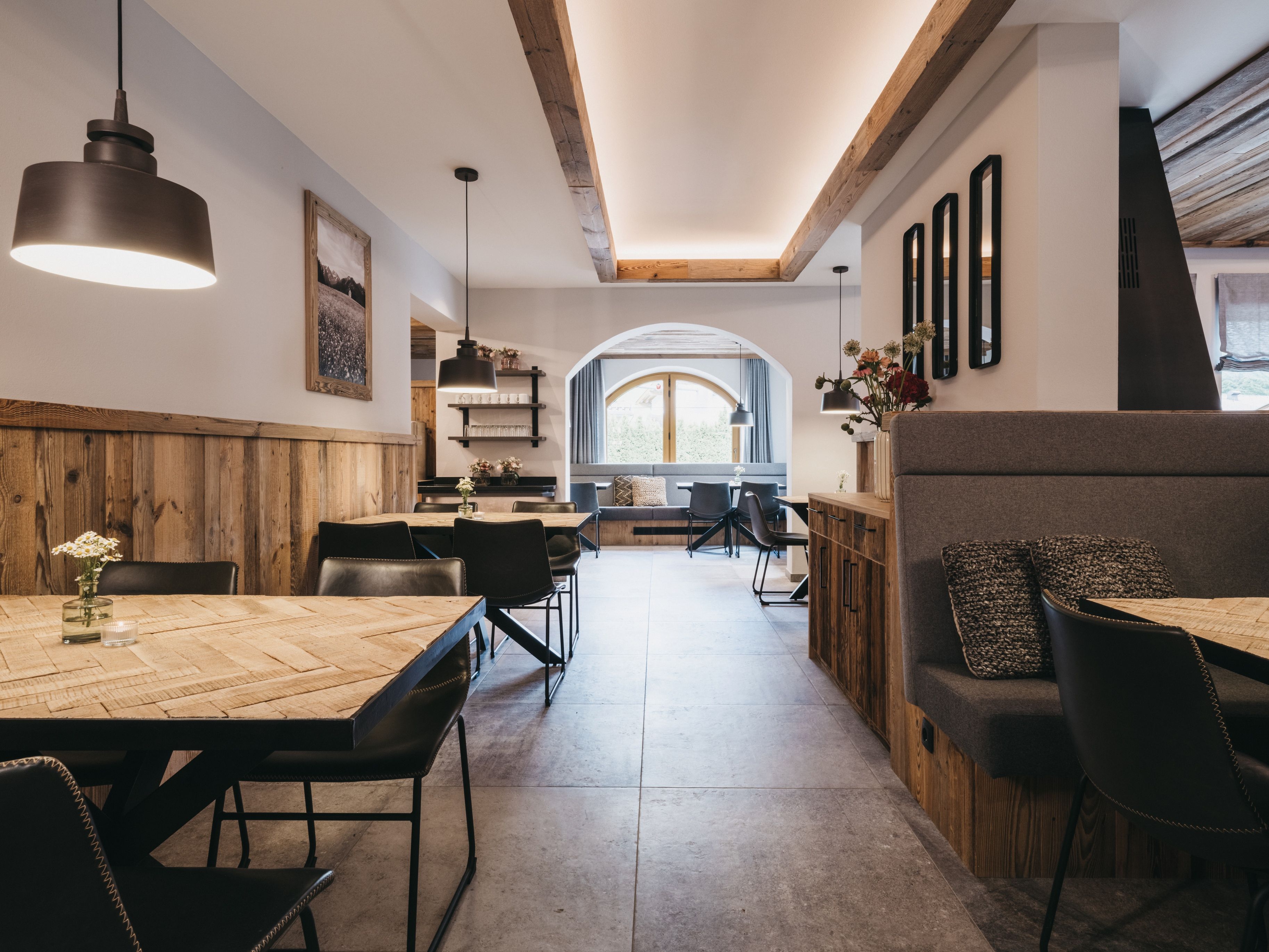 A modern dining area with a wooden table and chairs, pendant lighting, and a view through an arched window.