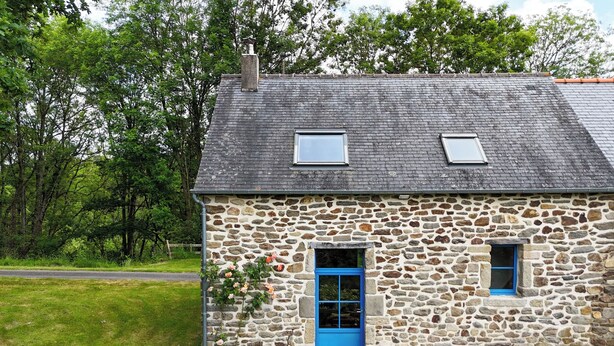 Una casa de piedra con puerta y ventanas azules, un pequeño espacio al aire libre con sillas y una mesa, y una cerca de madera al frente.