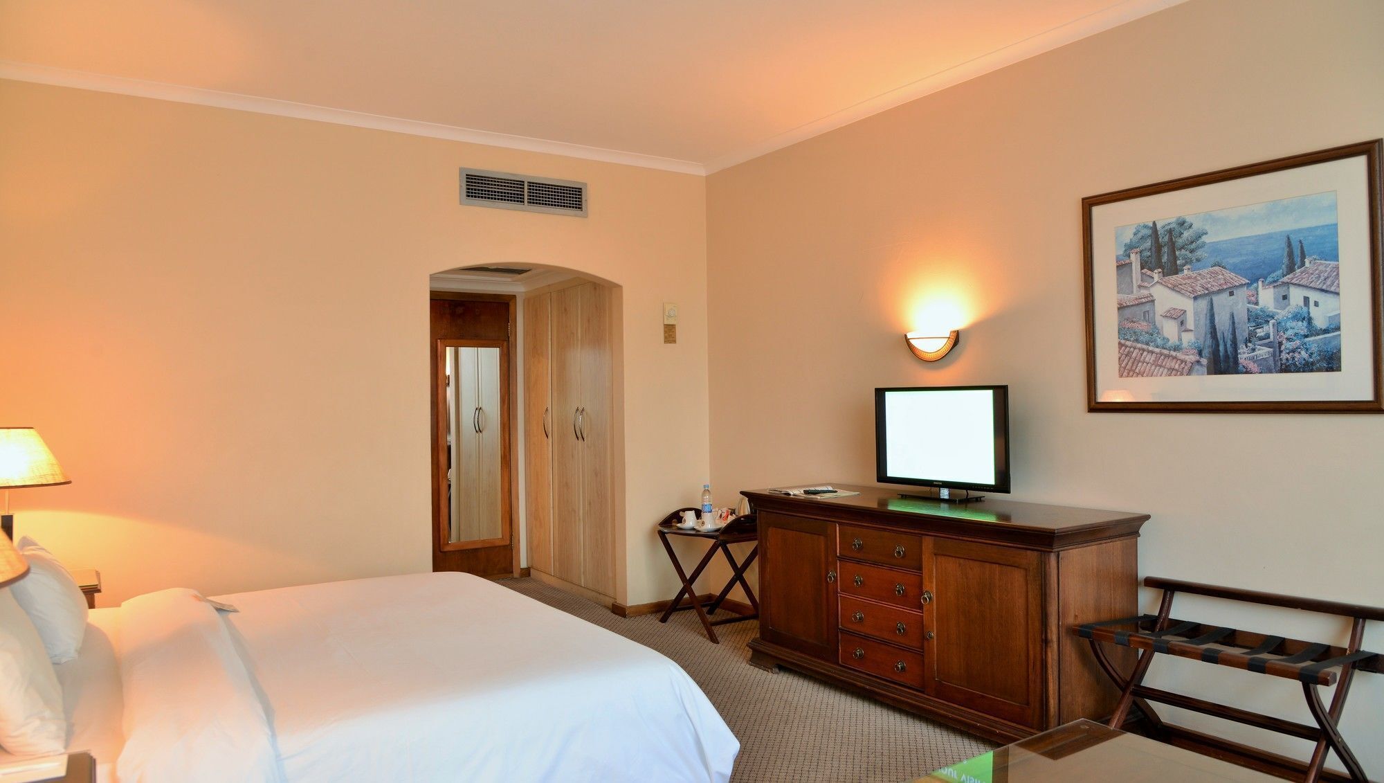 A hotel room with a bed, a desk with a computer, a chair, a lamp, a painting on the wall, and a door.
