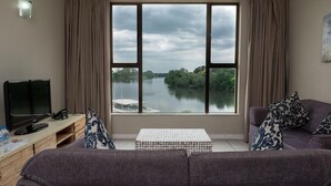 A living room with a large window offering a view of a river and greenery. There is a sofa, a coffee table, and a TV on a stand. - Riviera on Vaal Resort (Vereeniging)
