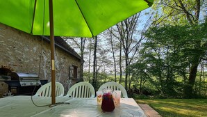 Une table avec un parasol vert, des chaises blanches et un barbecue à l’arrière-plan.