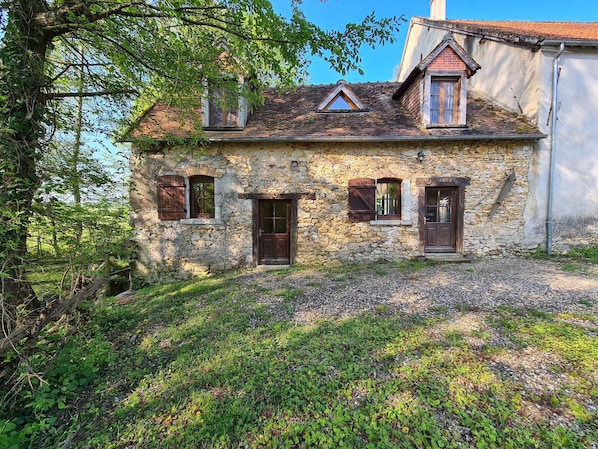 A stone house with a sloped roof, two windows with shutters, and a small porch area.