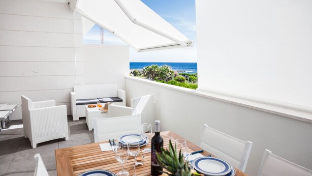 Een balkon met witte rieten meubels, een houten eettafel en uitzicht op zee.