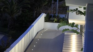 Een moderne buitenruimte met een betonnen pad, witte muren en een kleine tuin met planten en een bankje.