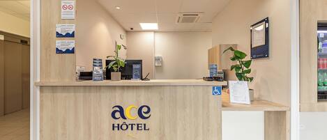 A hotel reception desk with a wooden finish, a sign indicating the hotel name, a wheelchair accessibility sign, and a digital screen displaying information.