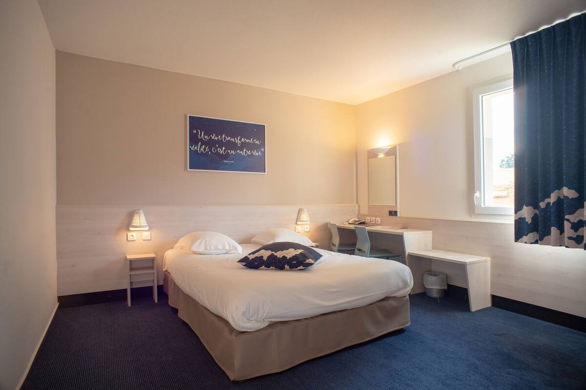 Une chambre d’hôtel avec un lit, des tables de chevet, un bureau, un miroir et une fenêtre avec des rideaux.