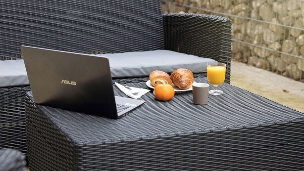 A wicker lounge chair with a laptop, croissants, an orange, and a glass of orange juice on a table.