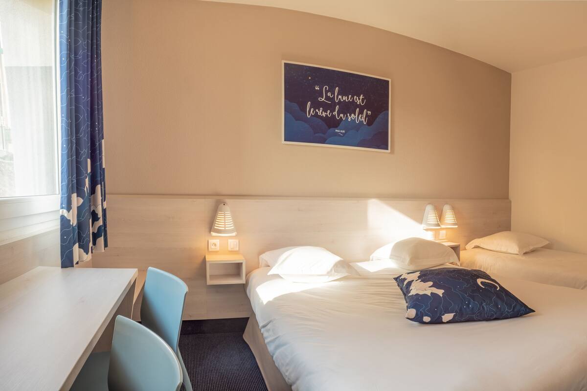 Une chambre d’hôtel avec deux lits, un bureau et une chaise. Il y a une affiche au mur et deux lampes de chevet.