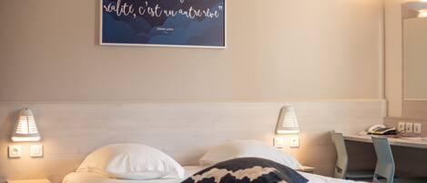 A hotel room with a bed, two bedside lamps, a desk, and a wall-mounted quote.