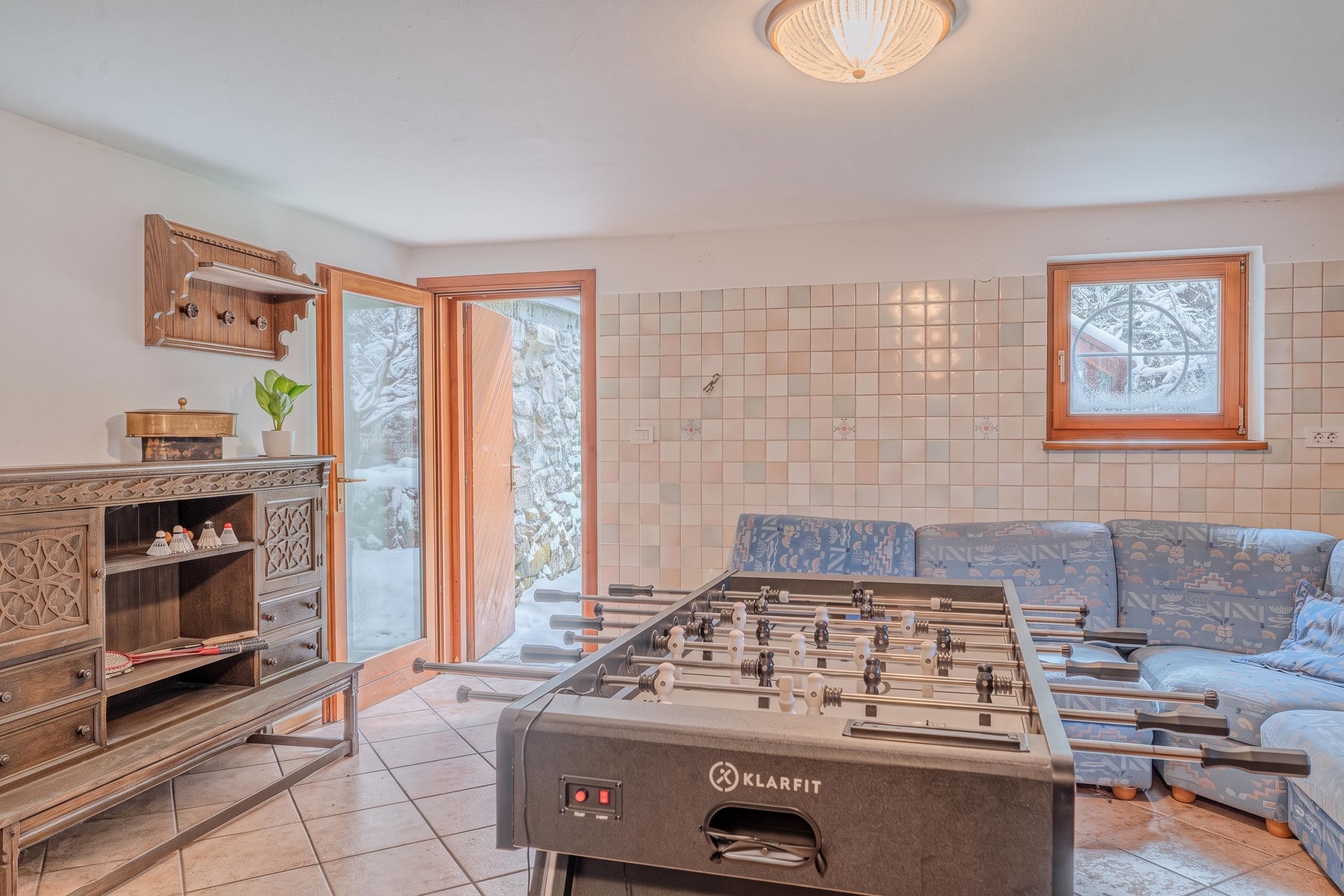 Una habitación con mesa de fútbol, sofá, aparador con objetos decorativos y ventana con vistas a árboles nevados.