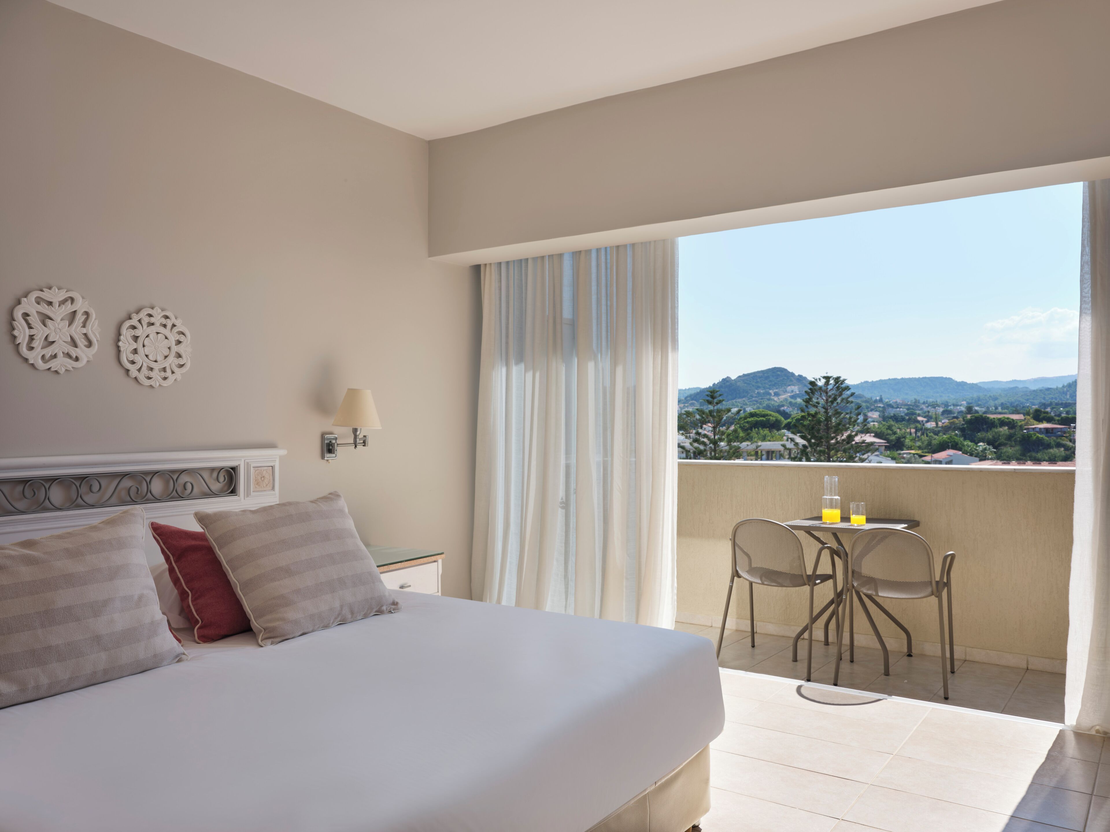 A hotel room with a bed, a nightstand, a lamp, a balcony with a table and chairs, and decorative wall art.