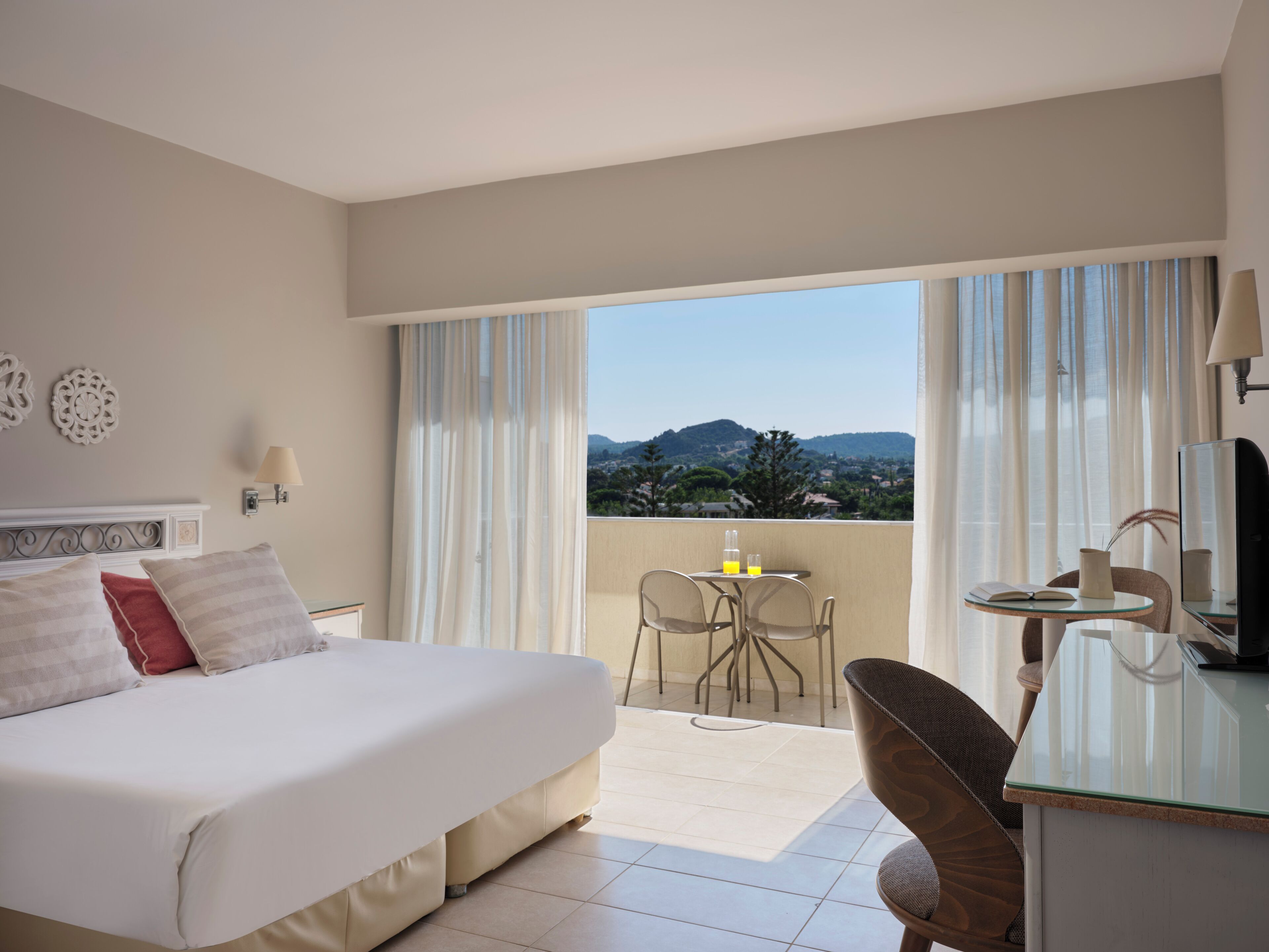 A hotel room with a bed, a desk, a chair, a table with a drink, and a balcony with a view of the mountains.