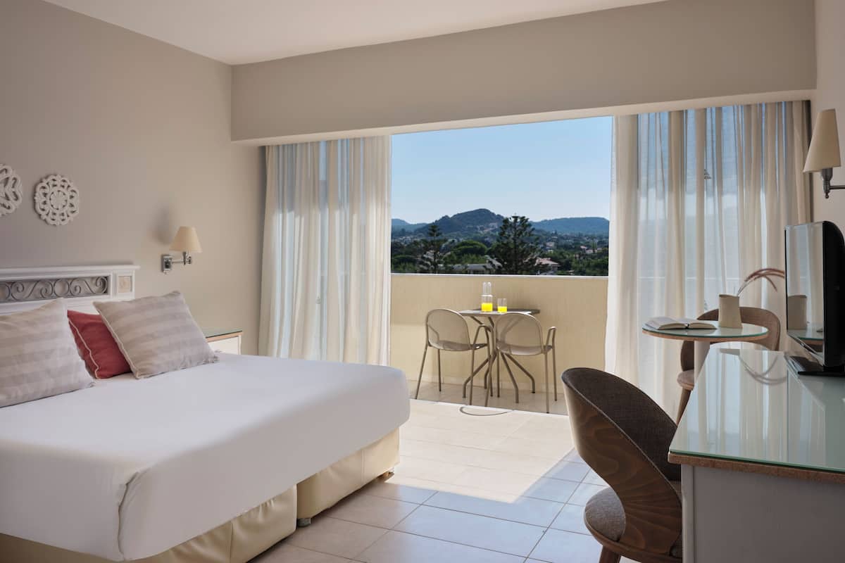Een hotelkamer met een bed, een bureau, een stoel, een tafel en een balkon met uitzicht op de bergen.