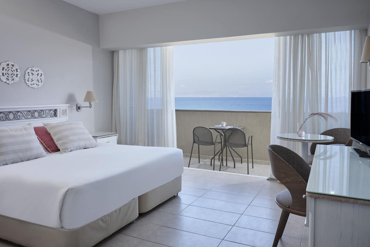Een hotelkamer met een groot bed, een bureau, een stoel en uitzicht op zee.