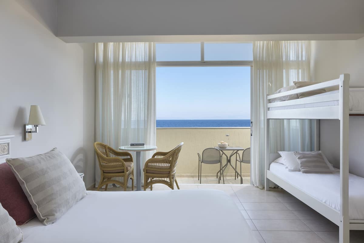 Een hotelkamer met een bed, een kleine tafel, twee stoelen, een balkon met uitzicht op zee, en een stapelbed.