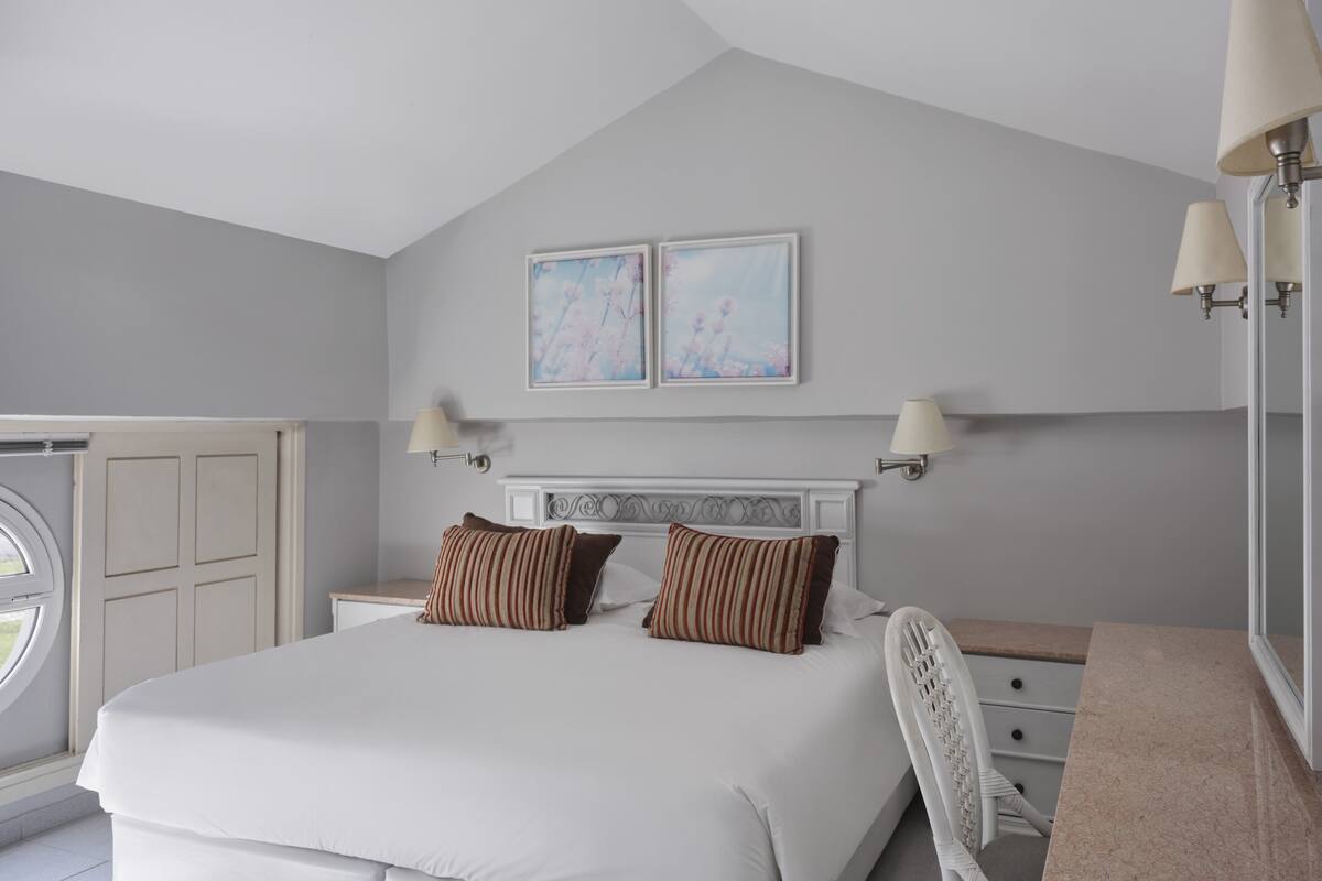 Een slaapkamer met een groot bed, twee wandlampen, een raam en twee ingelijste foto's aan de muur.