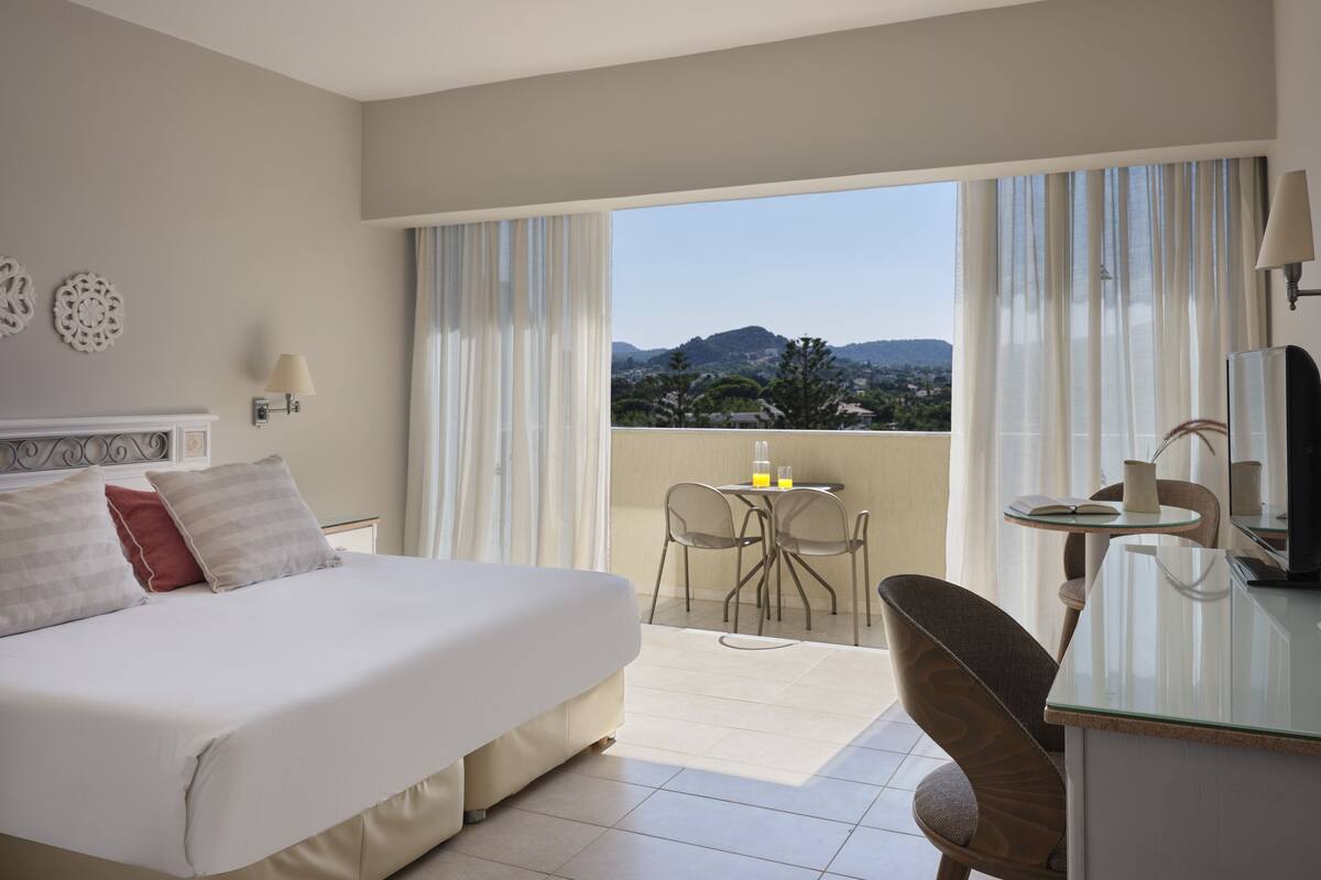 Een hotelkamer met een bed, een bureau, een stoel, een tafel en een balkon met uitzicht op de bergen.