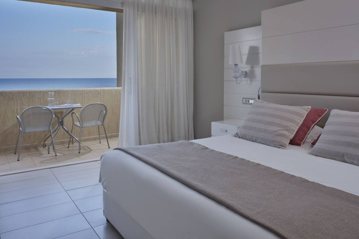 Een hotelkamer met een bed, nachtkastje, lamp, stoel en uitzicht op zee.