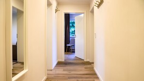 Een gang met houten vloer en witte wanden, die uitkomt in een kamer met een stoel en een raam.