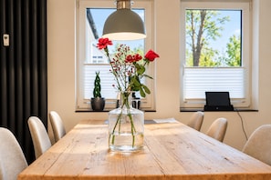 A wooden dining table with a clear glass vase containing red flowers and green stems, positioned in front of a window with blinds.