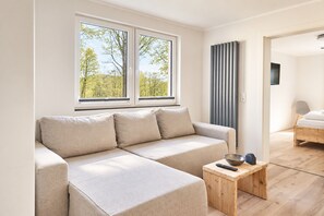 A modern living room with a sofa, a small wooden table, and a view of a forest through a window.