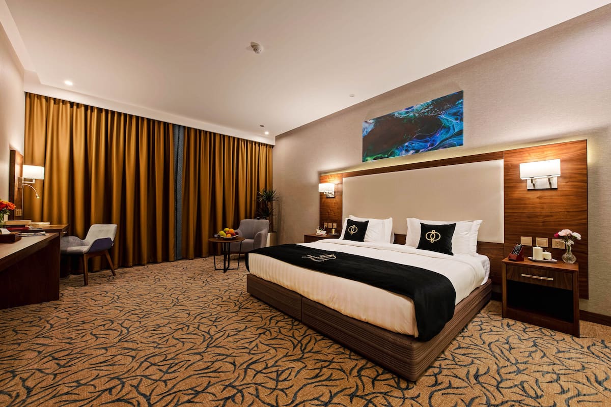 A hotel room with a large bed, a desk with a chair, a small table with fruit, and a painting on the wall.