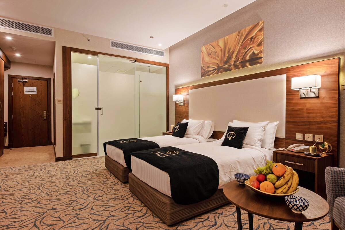 A hotel room with two beds, a glass shower door, a fruit bowl on the nightstand, and a wall-mounted lamp.