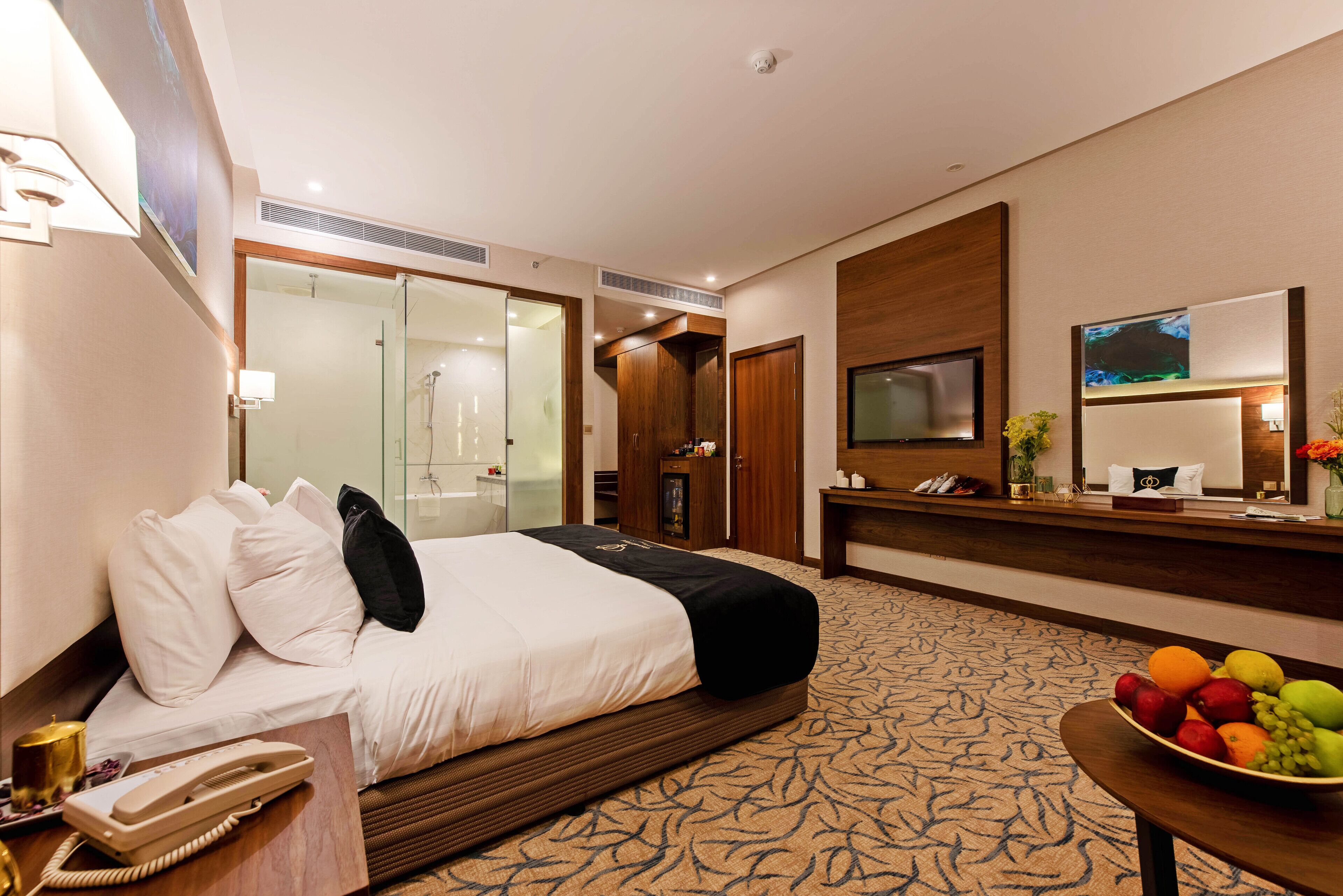 A hotel room with a bed, a desk with a telephone, a fruit bowl, a TV, and a bathroom visible through a glass door.