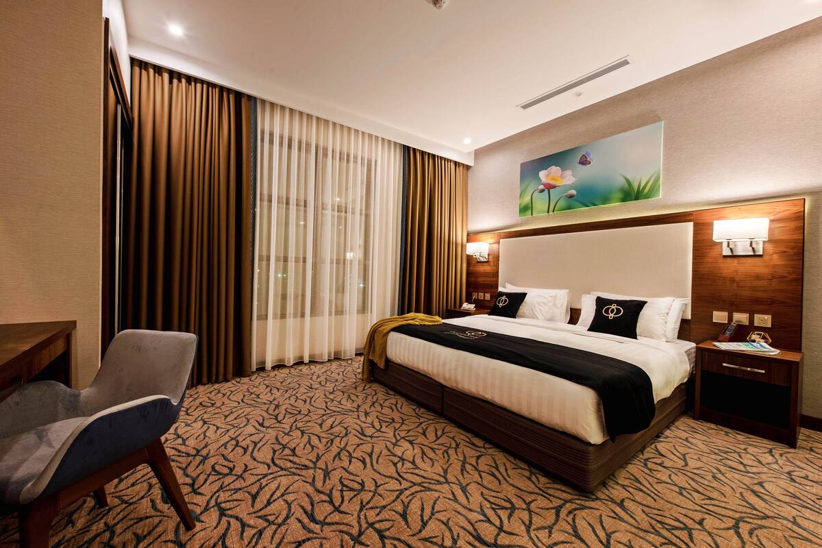 A hotel room with a large bed, a desk, and a chair. There is a painting on the wall and a window with curtains.