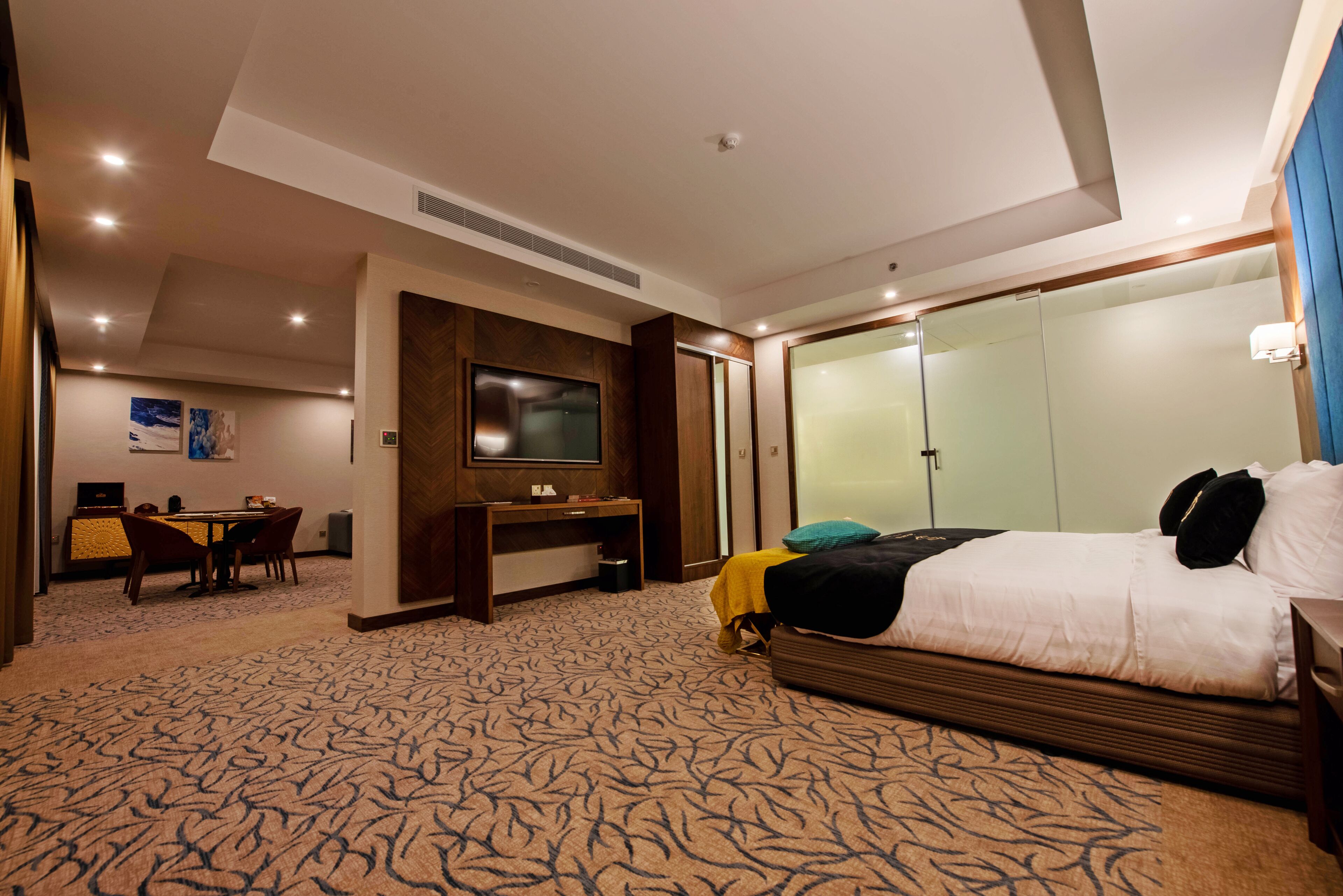 A hotel room with a large bed, a desk with a TV, a dining area, and a bathroom with a glass shower.