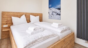 A double bed with white linens and pillows, a wooden headboard, a nightstand with a power outlet, and a wall-mounted radiator.