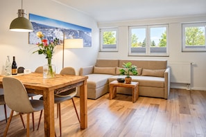 Ett modernt vardagsrum med ett matbord i trä, en soffa och ett soffbord. Det finns fönster med persienner, en lampa och en tavla på väggen.