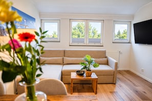 A living room with a sofa, a table, and a potted plant. There are windows with blinds and a flat-screen TV mounted on the wall. - Smart Resorts – Haus Opal – Holiday flat 405 with 1 bedroom, sleeps up to 4 (Winterberg)