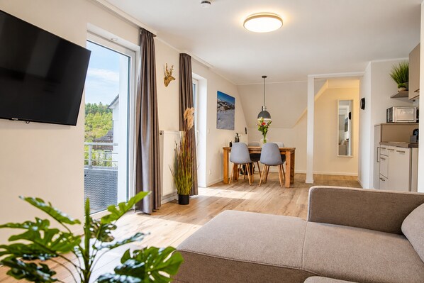 A modern living room with a flat-screen TV, a sofa, a dining area with a table and chairs, and a kitchen area with a microwave.