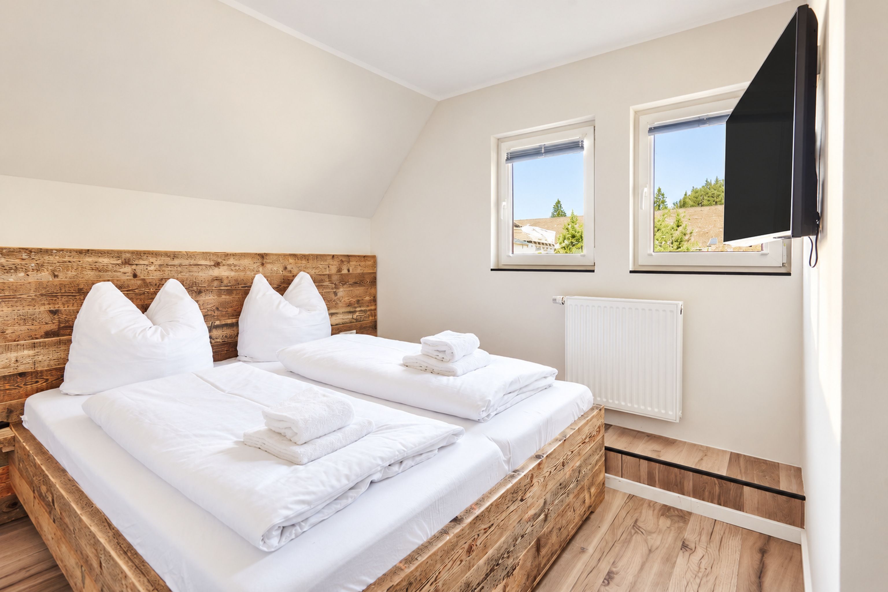 Ein Schlafzimmer mit einem hölzernen Kopfteil, zwei Betten, weißer Bettwäsche, einem Fernseher und einem Fenster mit Blick.