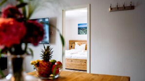 Une chambre avec un lit en bois, du linge de lit blanc, une petite table sur laquelle se trouve un panier de fruits, et un téléviseur fixé au mur.