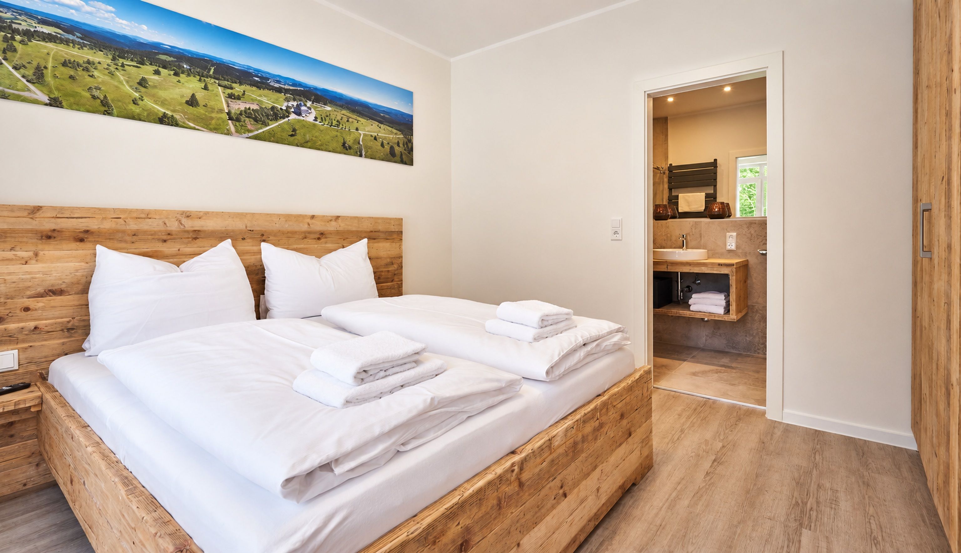 A hotel room with a wooden headboard, a large bed with white linens, a painting of a landscape on the wall, and a view into the bathroom.