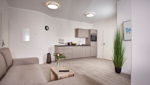 Een moderne woonkamer met een bank, een salontafel met een vaas en afstandsbediening, een keuken met kasten en apparatuur, en een potplant.