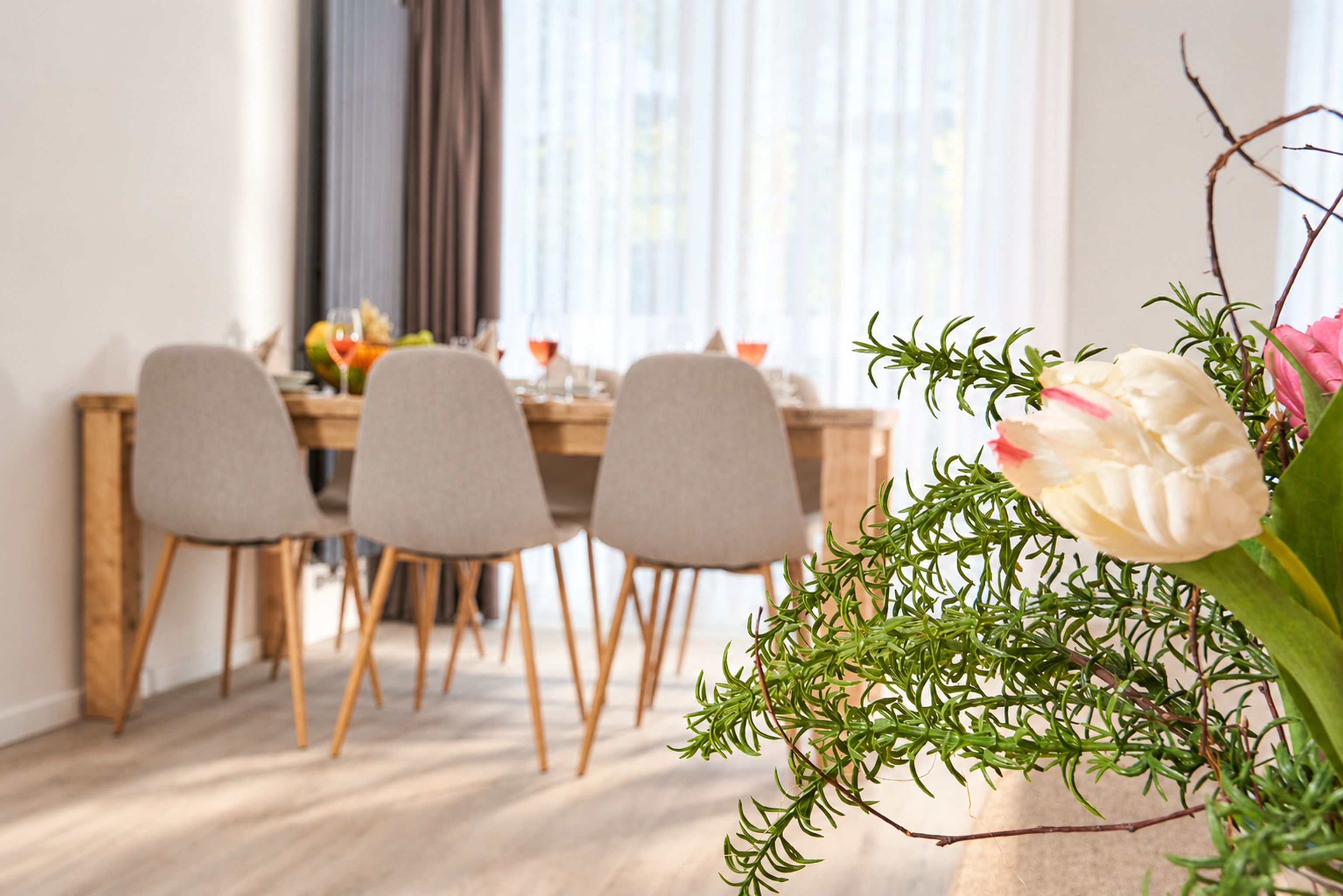 A dining area with a wooden table and four chairs, a vase with flowers, and a window with curtains.