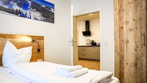 Een hotelkamer met een net opgemaakt bed, een houten hoofdeinde, een schilderij van een besneeerd landschap en uitzicht op de keuken.