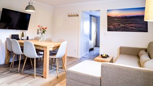 A modern living room with a wooden dining table, grey upholstered chairs, a grey sofa, and a flat-screen TV mounted on the wall.