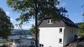 A two-story white building with a sloped roof, surrounded by trees and a view of solar panels in the background.
