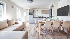 A modern living room with a sofa, dining table, and chairs. A kitchen with an oven and a TV is visible in the background.