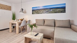 A modern living room with a sofa, a coffee table, a dining table with chairs, a hanging light, and a landscape painting on the wall. - Smart Resorts - Haus Azur - Ferienwohnung 808, Max. 4 Personen (Winterberg)