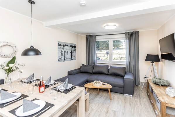 Ein modernes Wohnzimmer mit einem grauen Sofa, einem Esstisch für vier Personen und einem großen Fenster mit Vorhängen.