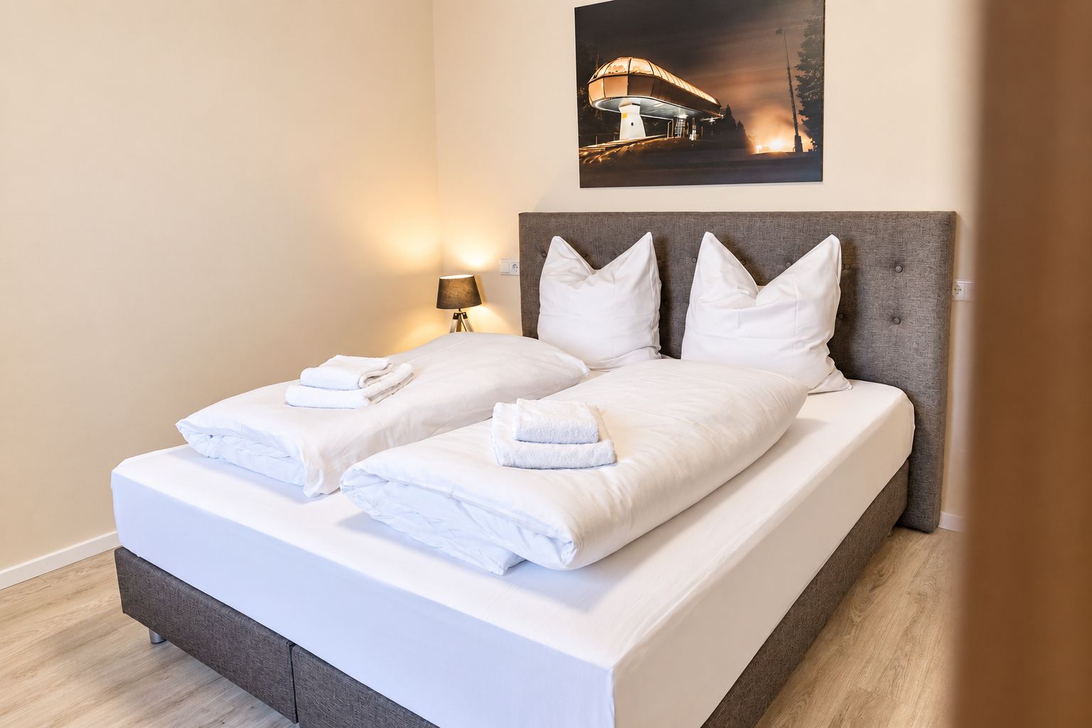 A double bed with white linens and towels, a grey upholstered headboard, and a wall-mounted lamp.