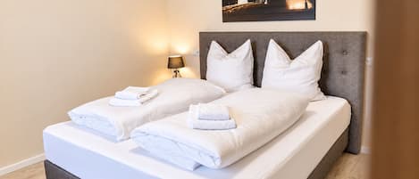 Cama doble con ropa de cama y toallas blancas, cabecera tapizada de gris y lámpara fijada a la pared.