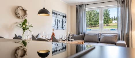 A modern living room with a dining area, a large window with curtains, and a wall-mounted artwork.
