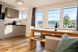 A modern kitchen with a dining area, wooden furniture, and a view of a forest through large windows.