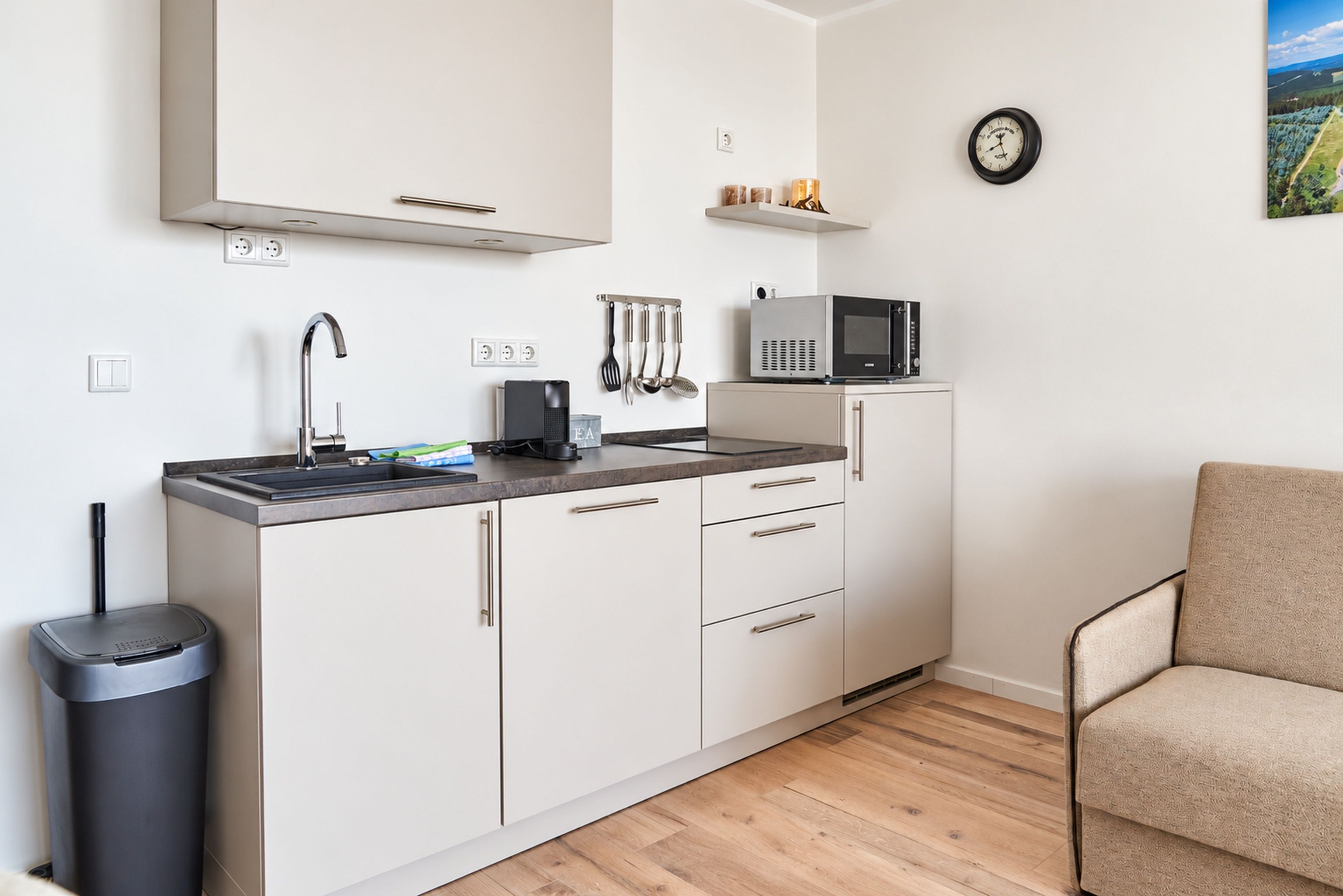 A compact kitchenette with a microwave, kettle, and utensils on the counter, a trash bin, and a chair in the corner.